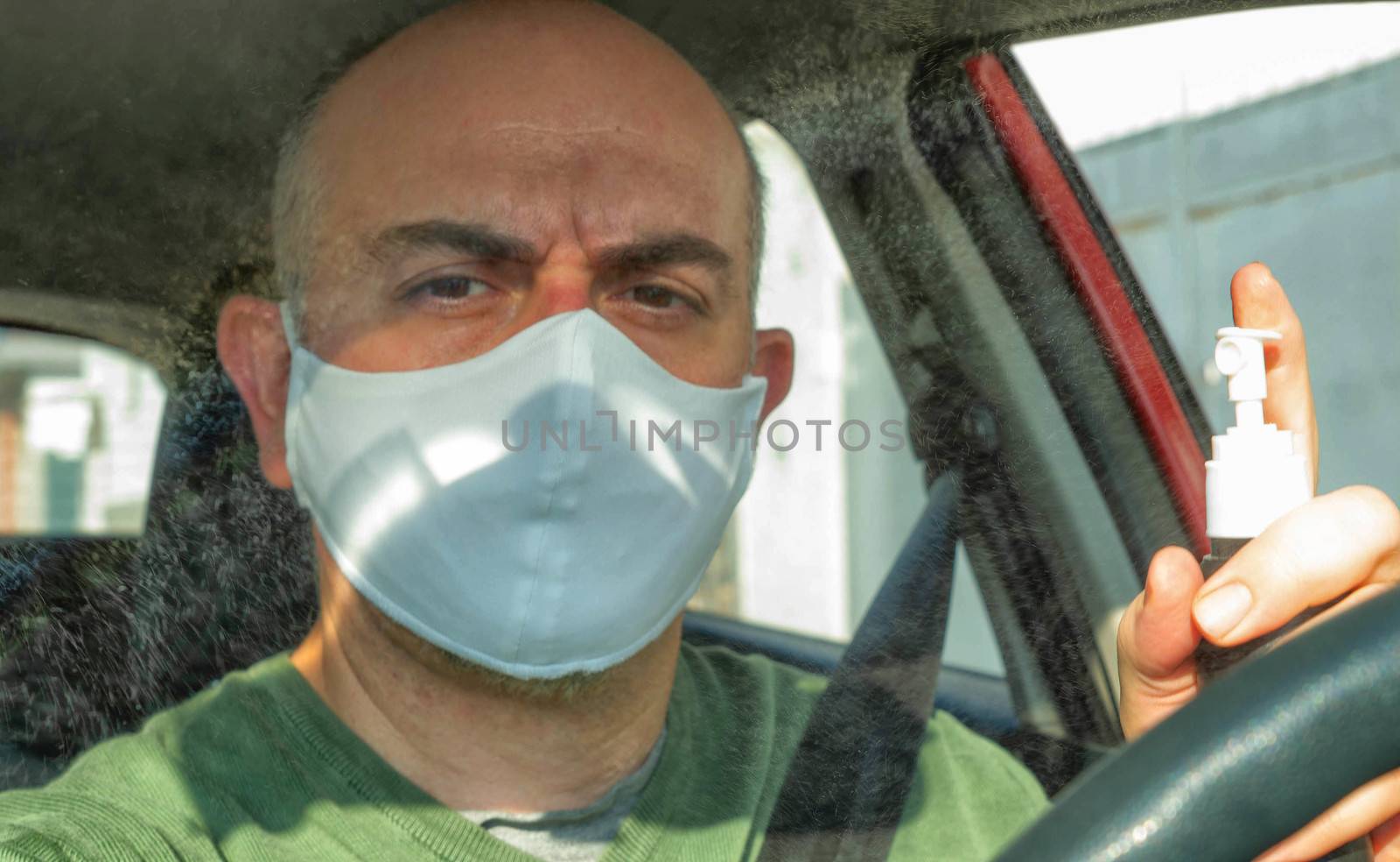 Turin, Piedmont, Italy. April 2020. Portrait of a Caucasian man driving the car wearing a white mask to avoid contagion. Mist alcohol to disinfect surfaces: droplets shine suspended in the air.