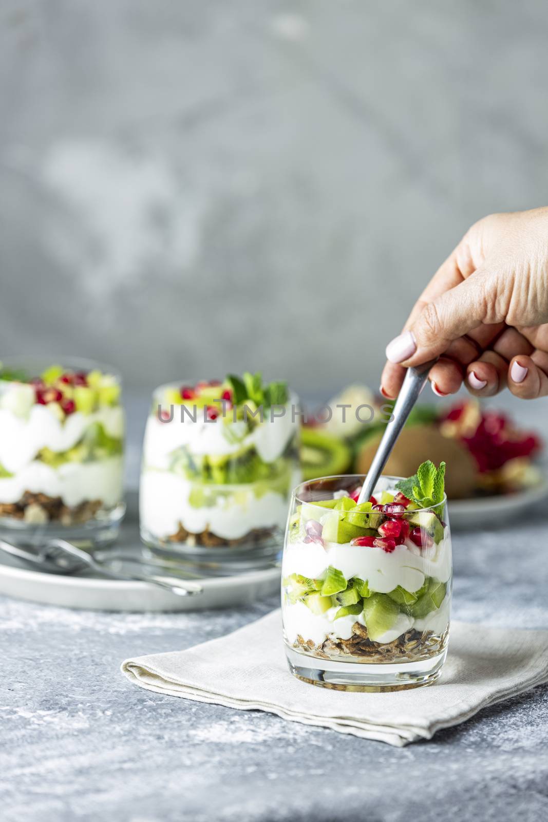 Person woman hand inserting spoon in the breakfast jar with granola, yogurt and kiwi fruits on gray surface. Delicious food, traditional american snack.