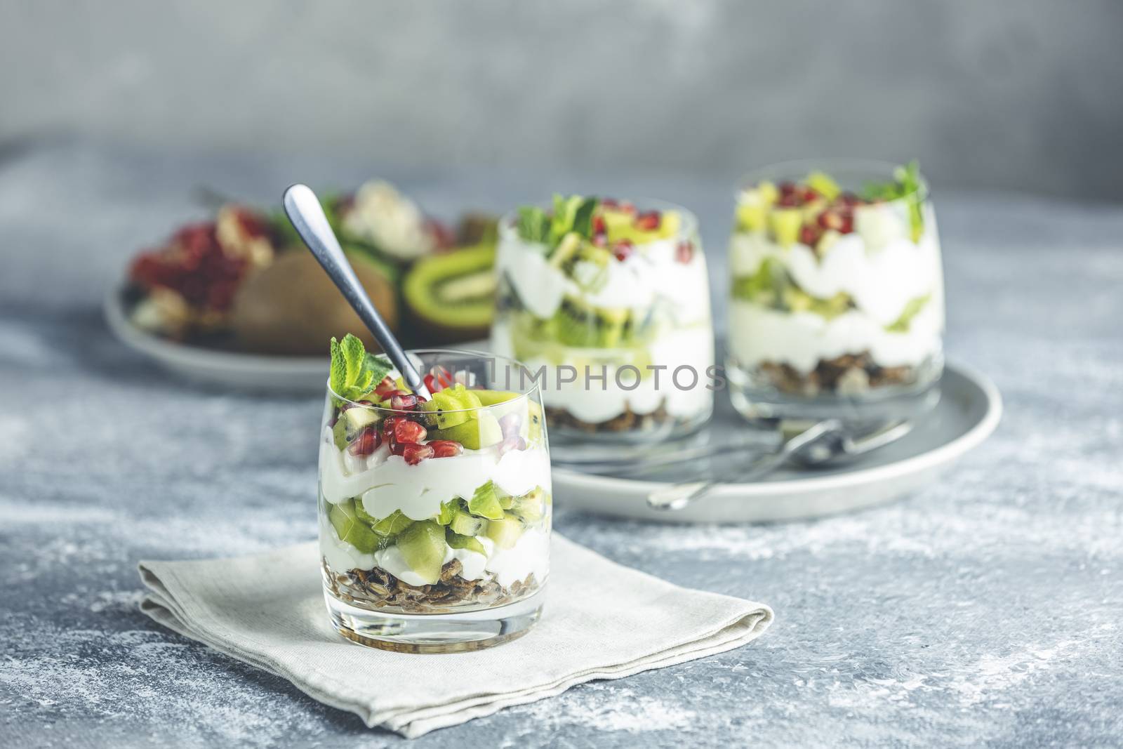 Glass jar of kiwi parfait dessert in glass with ingredients with spoon on gray napkin. Yogurt, granola and fruits. Healthy snack or breakfast. Light gray concrete surface.