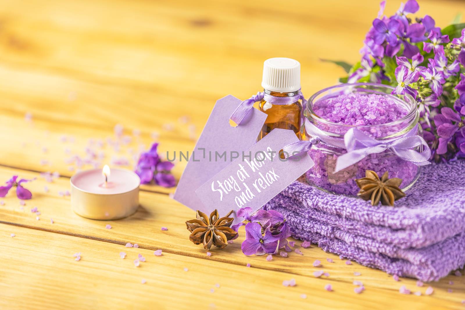 Stay at home and relax text phrase on label sticker. Spa still life with violet oil, towel and perfumed candle on natural wood table surface. Quarantine coronavirus life concept.