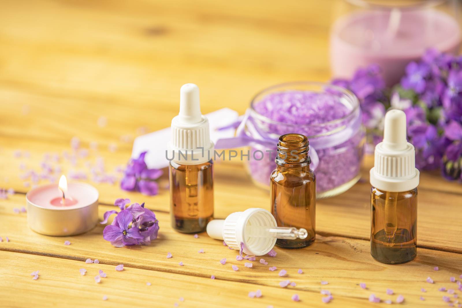 Spa still life with violet oil, violaceous bath salt in glass jar and perfumed candle on natural wood table surface.