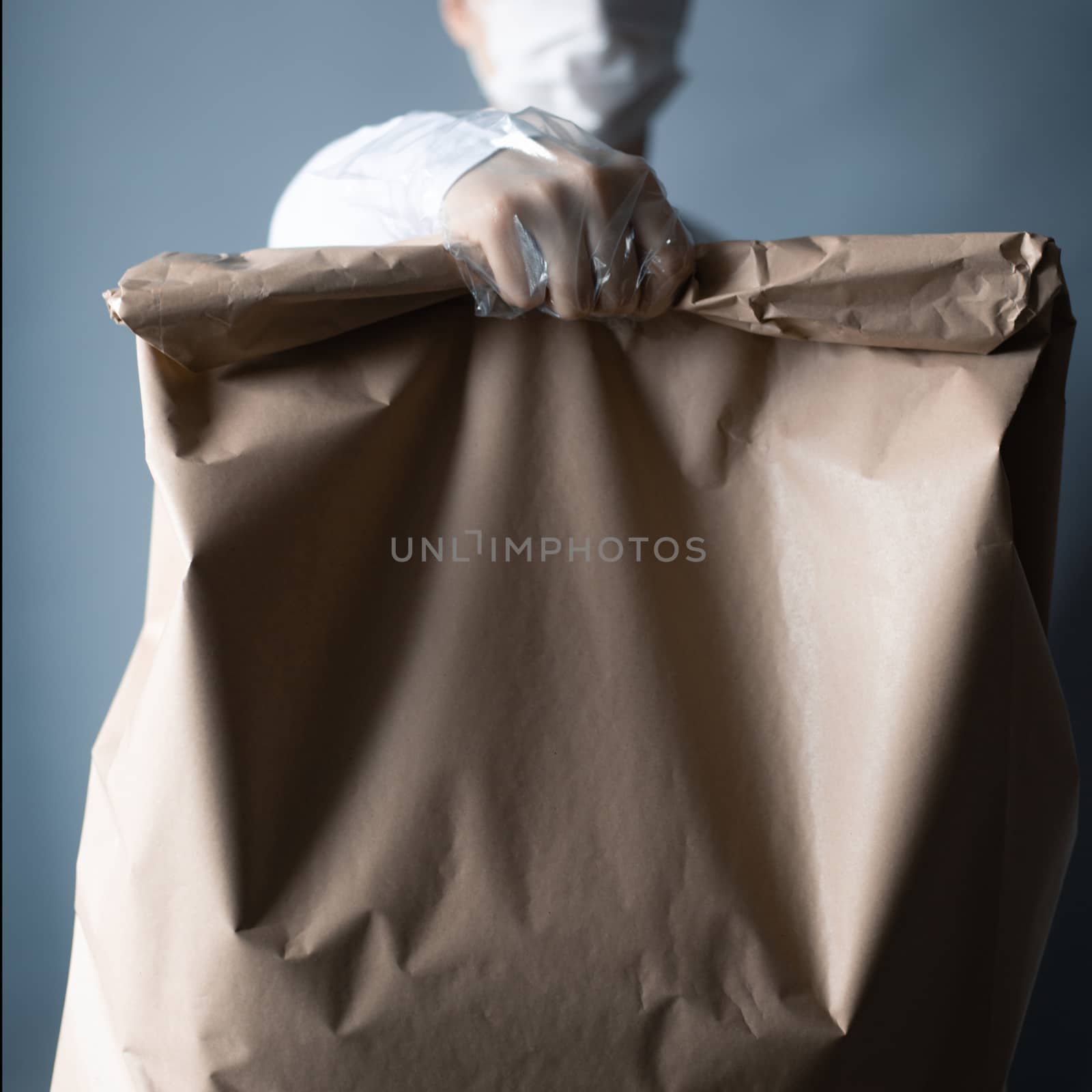 Safe food or goods delivery. Young courier delivering grocery brown eco paper bag order to the home of customer with mask and gloves during the coronavirus pandemic. White clothes gray background copy space for text