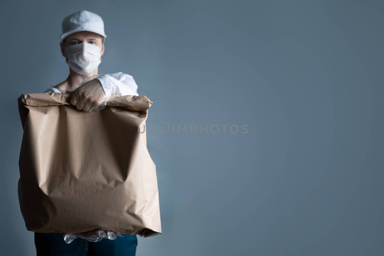 Safe food or goods delivery. Young courier delivering grocery brown eco paper bag order to the home of customer with mask and gloves during the coronavirus pandemic. White clothes gray background copy space for text