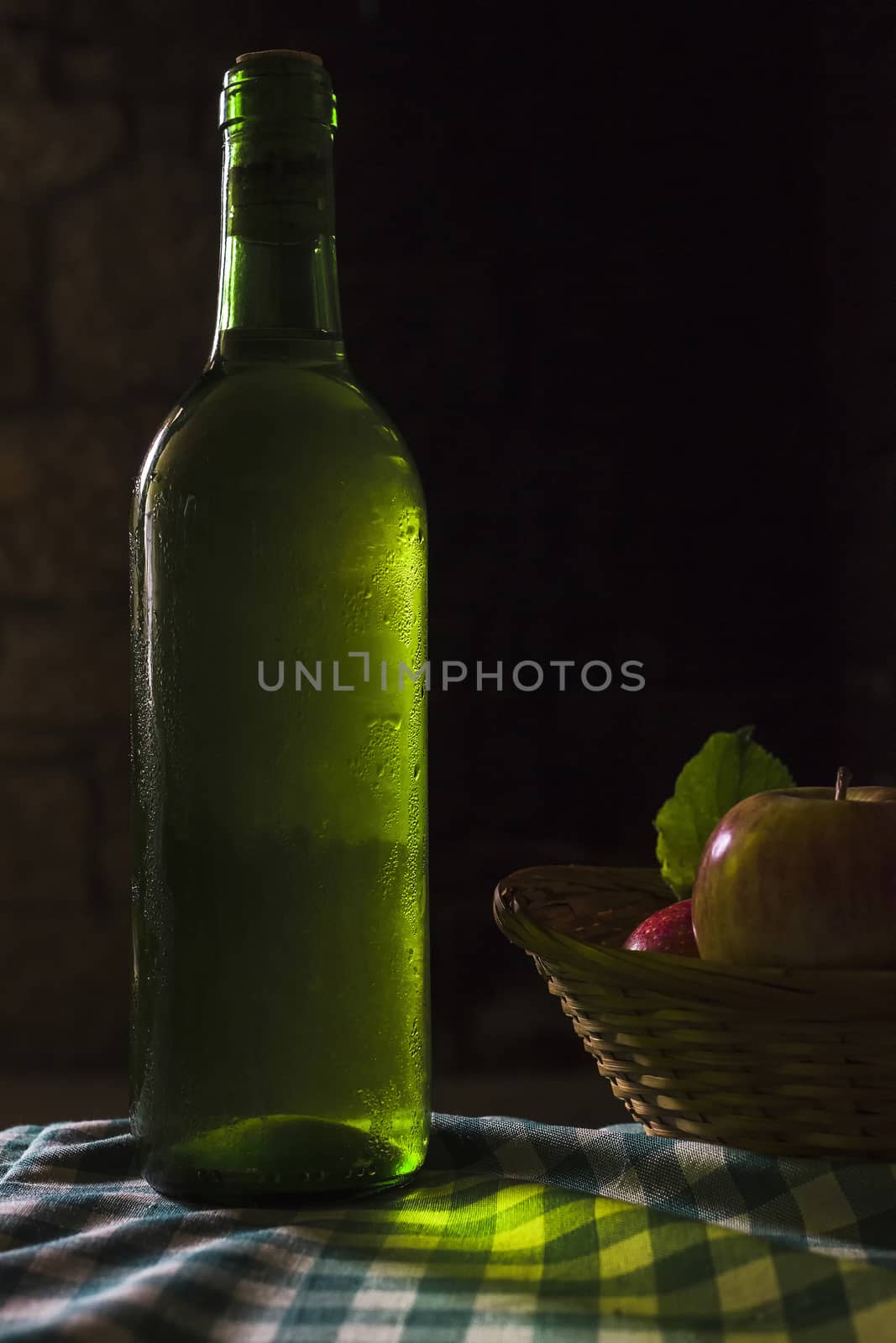 Green bottle of white wine with condensation drops and basket of apples on a dark background