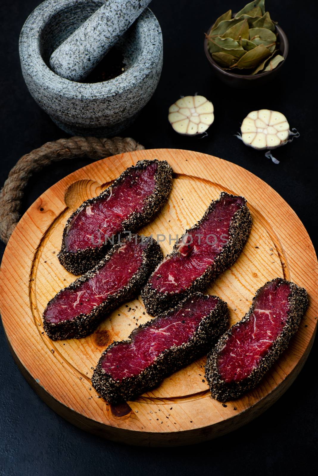 Stone mortar bowl with pestle, garlic, bay leaf and wooden cutting board with raw pepper steaks on stone surface