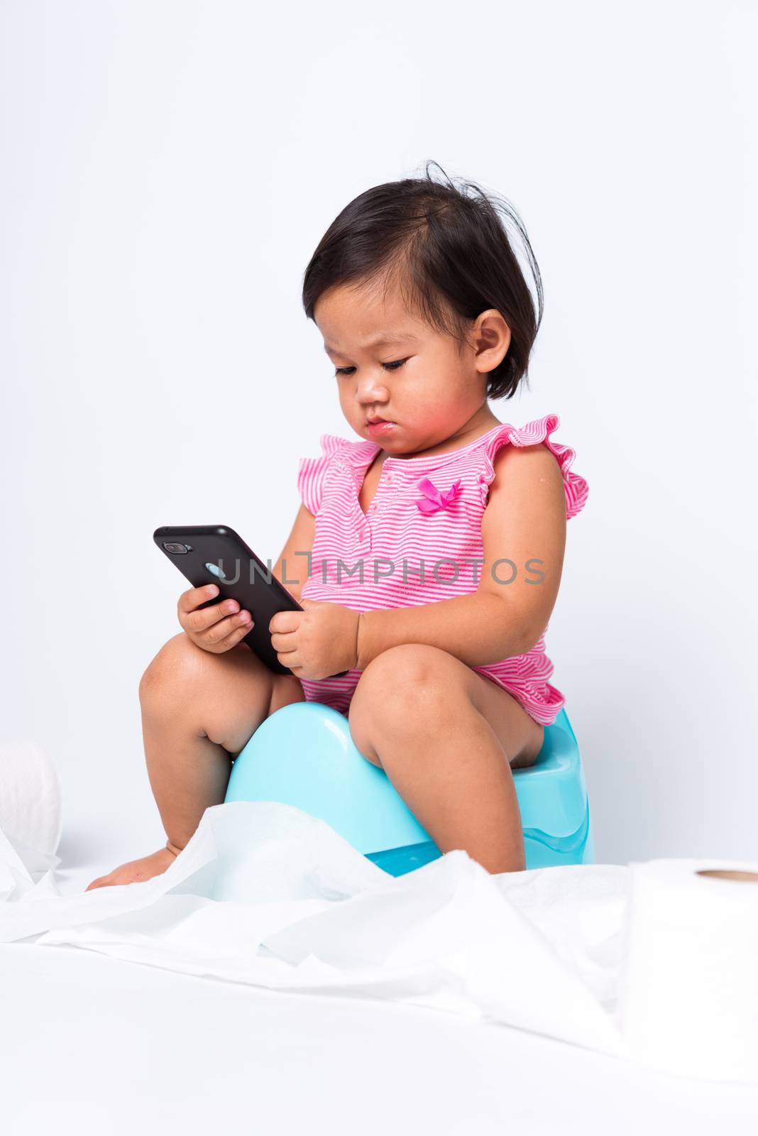 Asian little cute baby child girl education training to sitting on blue chamber pot or potty and play smart mobile phone with toilet paper rolls, studio shot isolated on white background, wc toilet