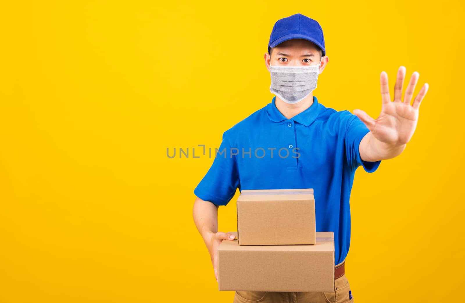 Asian young delivery worker man in blue t-shirt and cap uniform wearing face mask protective hold boxes he raise hand to stop sign under coronavirus or COVID-19, studio shot isolated yellow background