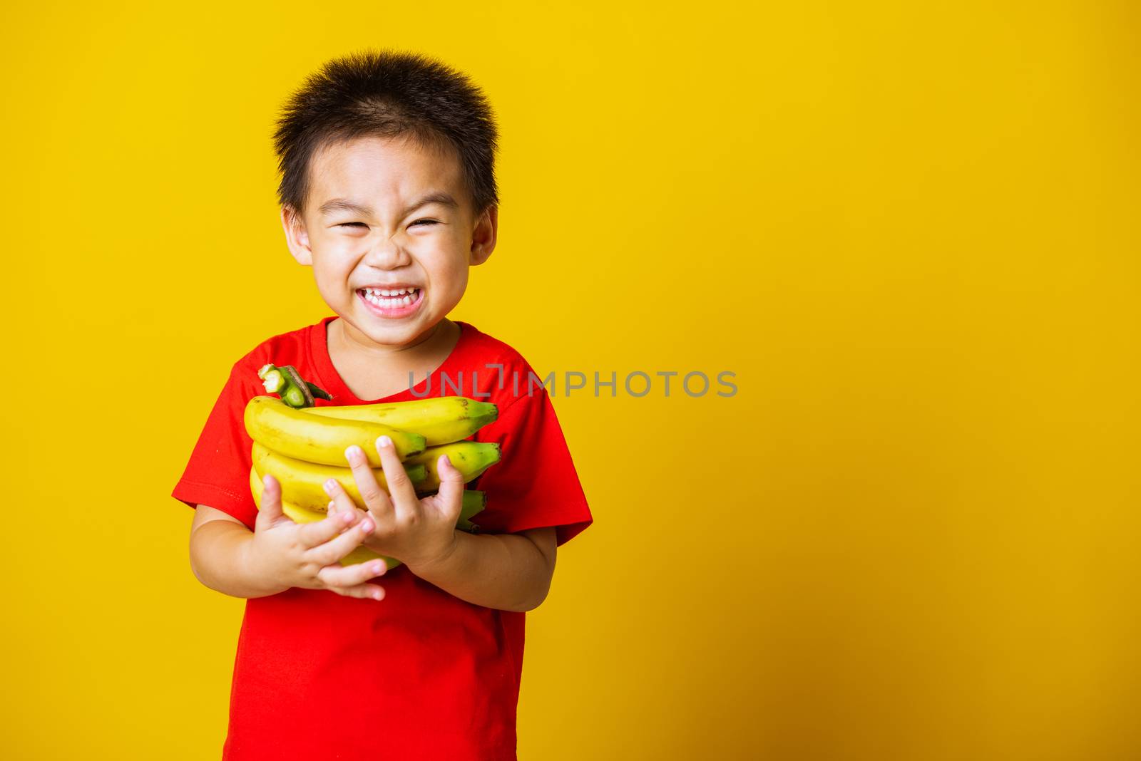 Happy portrait Asian child or kid cute little boy attractive smile wearing red t-shirt playing holds bananas comb fruit, studio shot isolated on yellow background