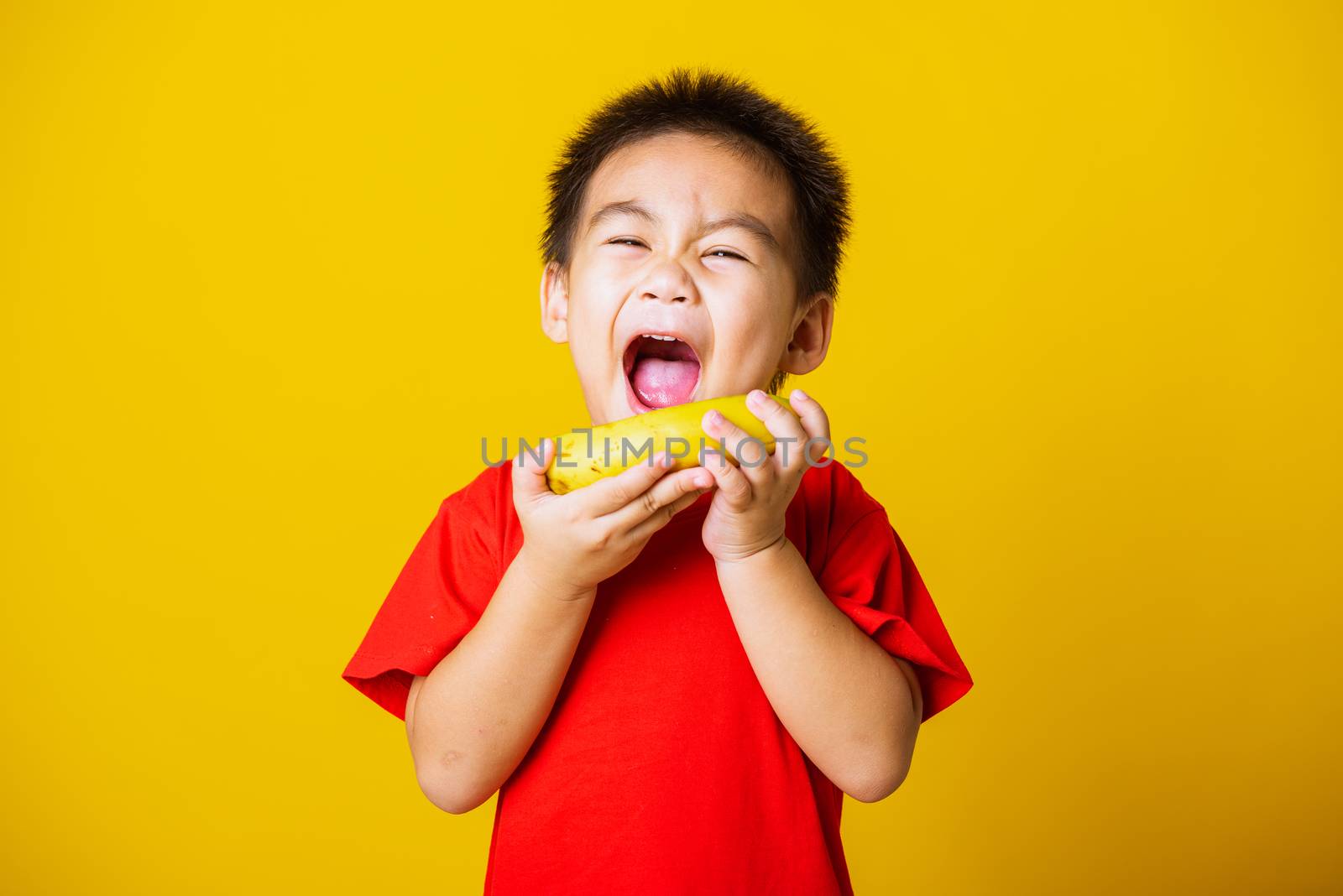 Happy portrait Asian child or kid cute little boy attractive smile wearing red t-shirt playing holds banana fruit, studio shot isolated on yellow background