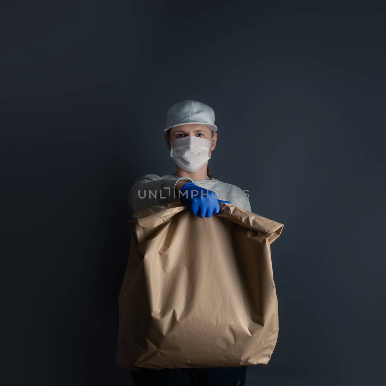 Safe food or goods delivery. Young courier delivering grocery big brown eco paper bag order to the home of customer with mask and gloves during the coronavirus pandemic. Gray background copy space for text