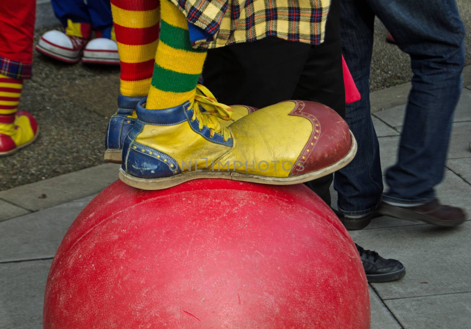 Close up view of a clown wearing outsize, colourful shoes while balancing on a large ball.
