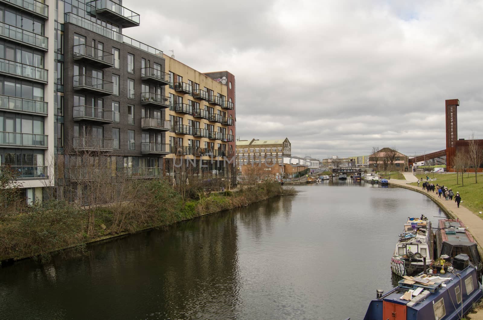 Walkers enjoying the views on a cloudy March day along the Lee Navigation, a canalised part of the River Lea running between Hackney Wick and the Queen Elizabeth Park in Newham, East London.  The Olympic Park Energy Centre is on the right hand side.