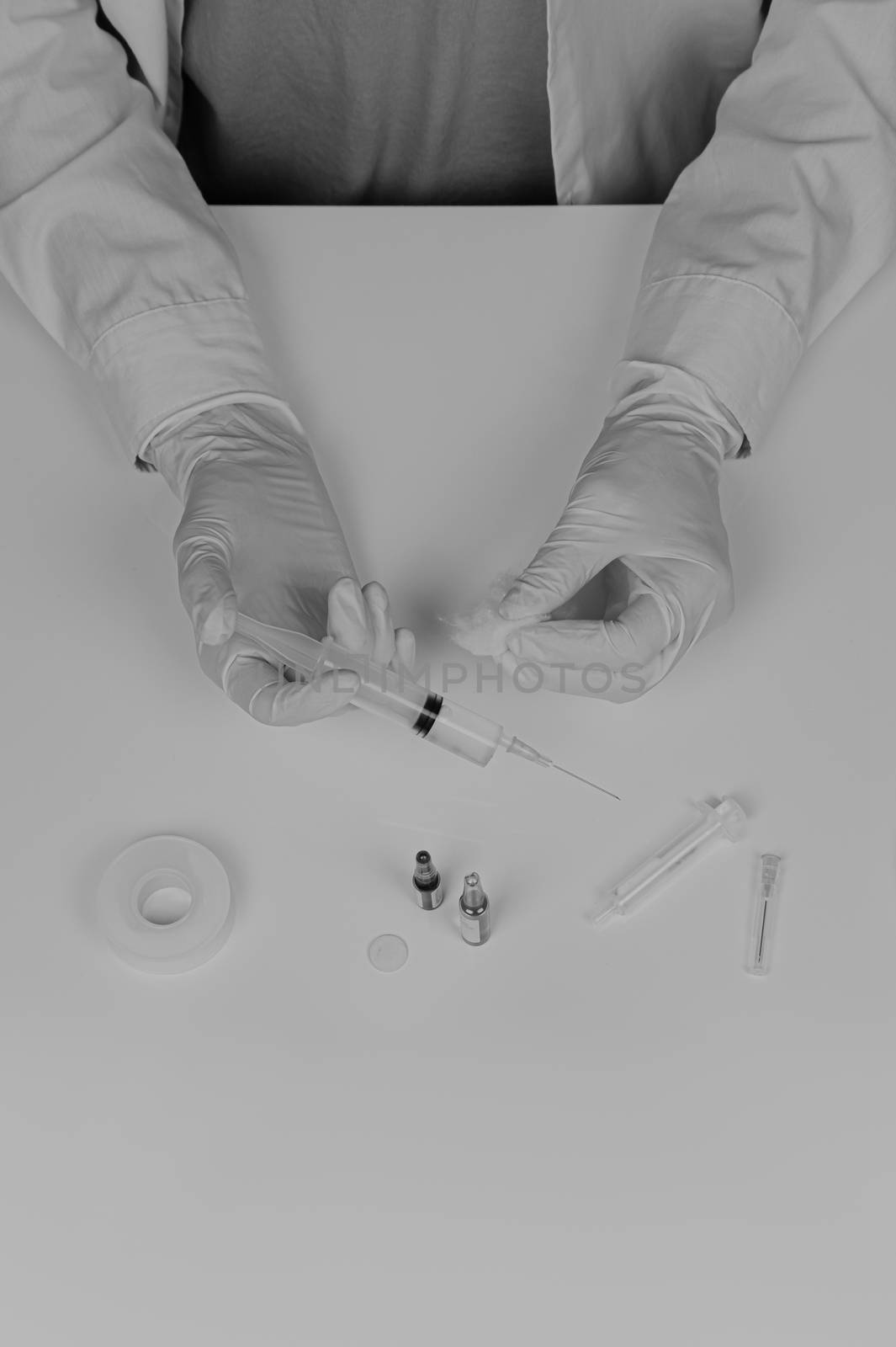MD preparing injection, faded monochrome style. Top view of medical doctor hands in gloves and lab coat holding syringe on white table with medical accessories