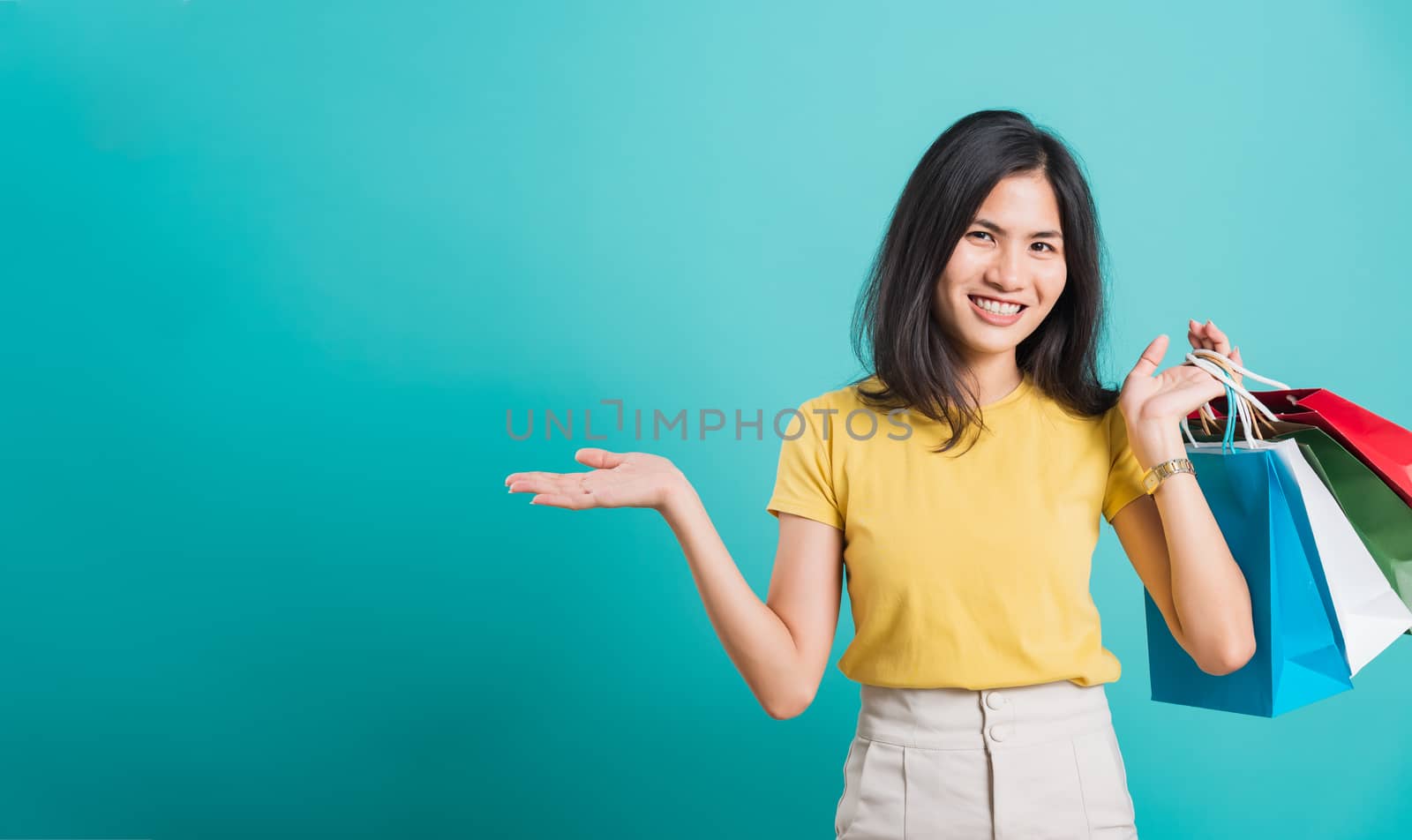 Portrait happy Asian beautiful young woman smile white teeth standing wear yellow t-shirt, She holding shopping bags show hand something empty and looking to camera, studio shot on blue background