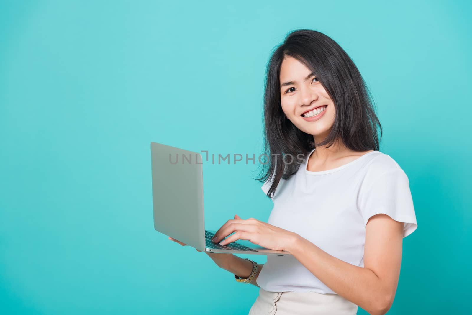 Portrait happy Asian beautiful young woman smile white teeth standing wear white t-shirt, She holding and using a laptop computer, studio shot on blue background with copy space for text