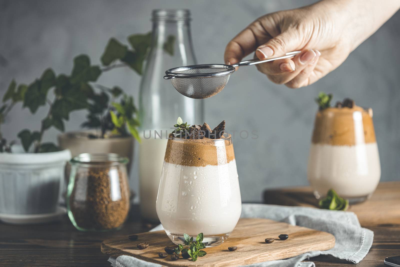 Woman hand sifting cocoa powder by sieve over glass of iced frothy drink Dalgona Coffee. Trend korean drink latte espresso with coffee. Dark rustic style.