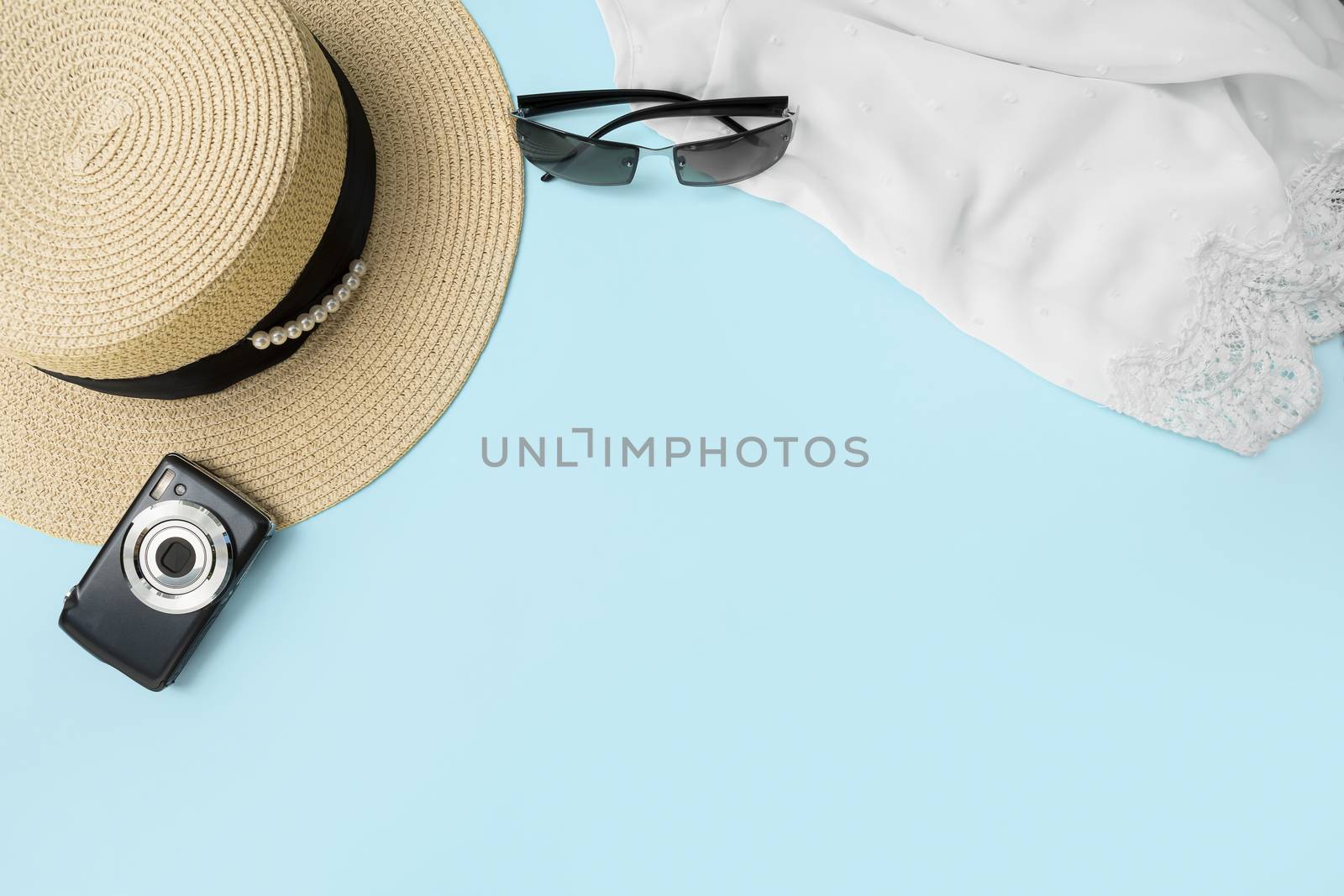 Summer vacation concept with light blue background. Straw hat decorated with black ribbon and pearls. Next to it is a black compact camera, sunglasses and a white women's lacy top. Space for the copy.