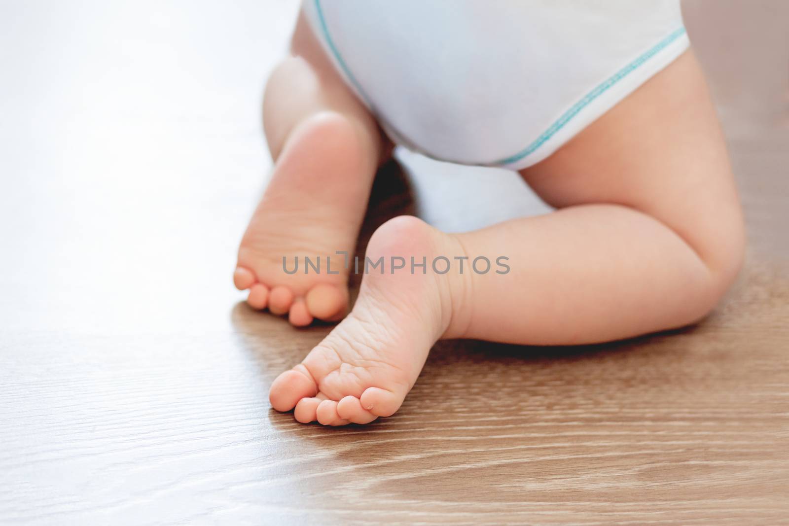 Baby's feet on wooden floor. Bare heels of little child wearing white bodysuit and diaper. Cozy morning bedtime at home.