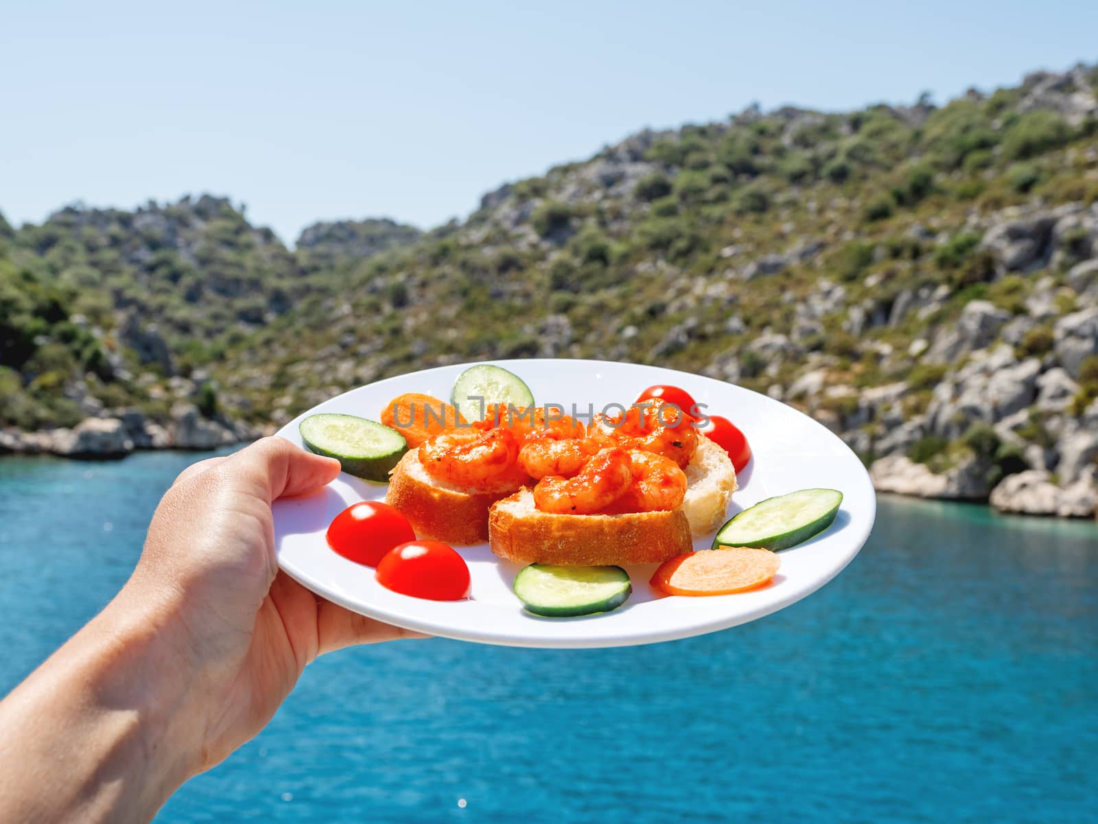 Mediterranean cuisine - freshly cooked shrimps on bread with vegetables. Hand with plate on beautiful sea lagoon background. Seafood with cucumbers and tomatoes.