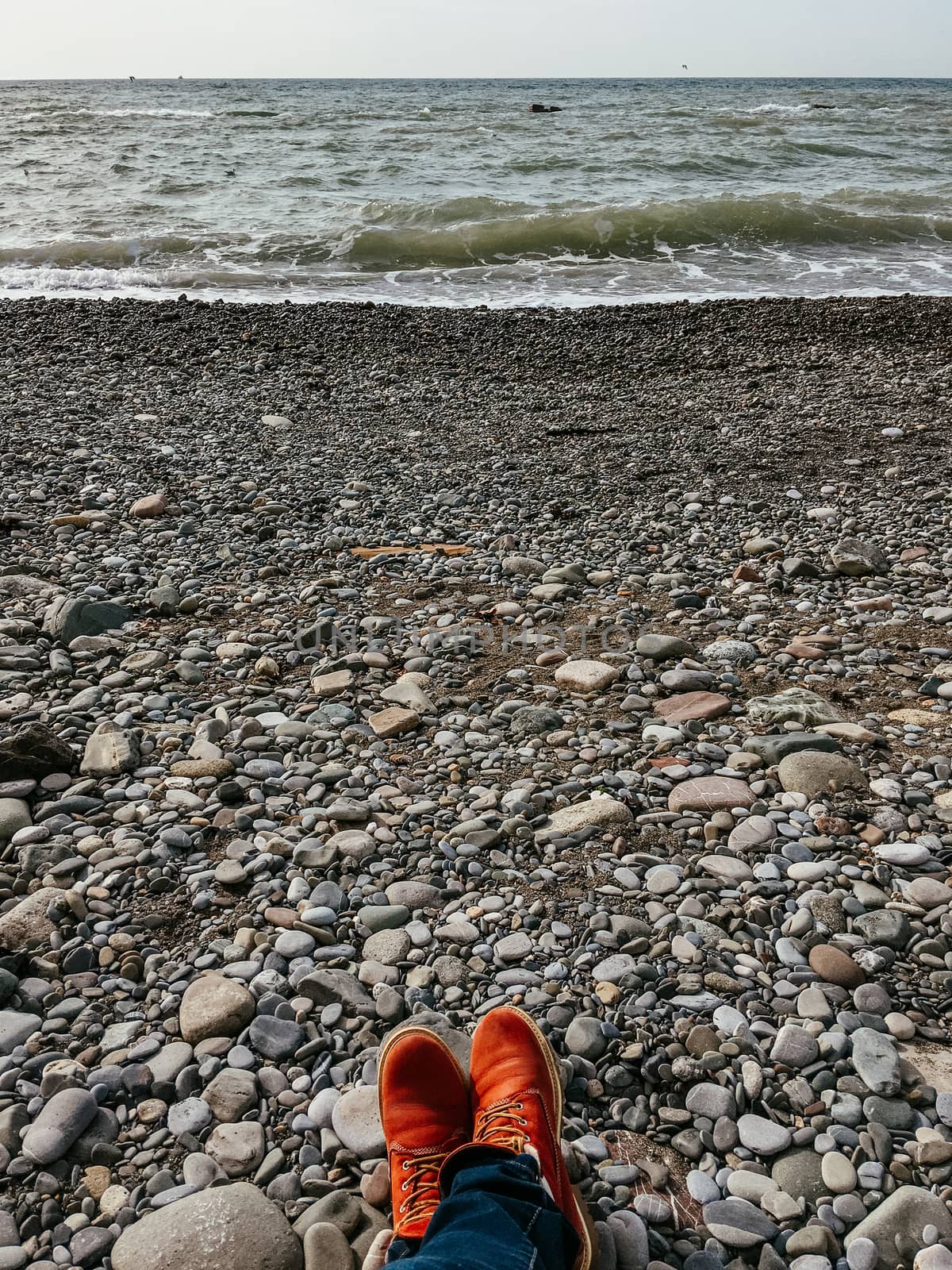 Feet in red boots on seaside. View on seascape from rocky beach. Travel concept. Man or woman in hiking shoes is relaxing outdoors.