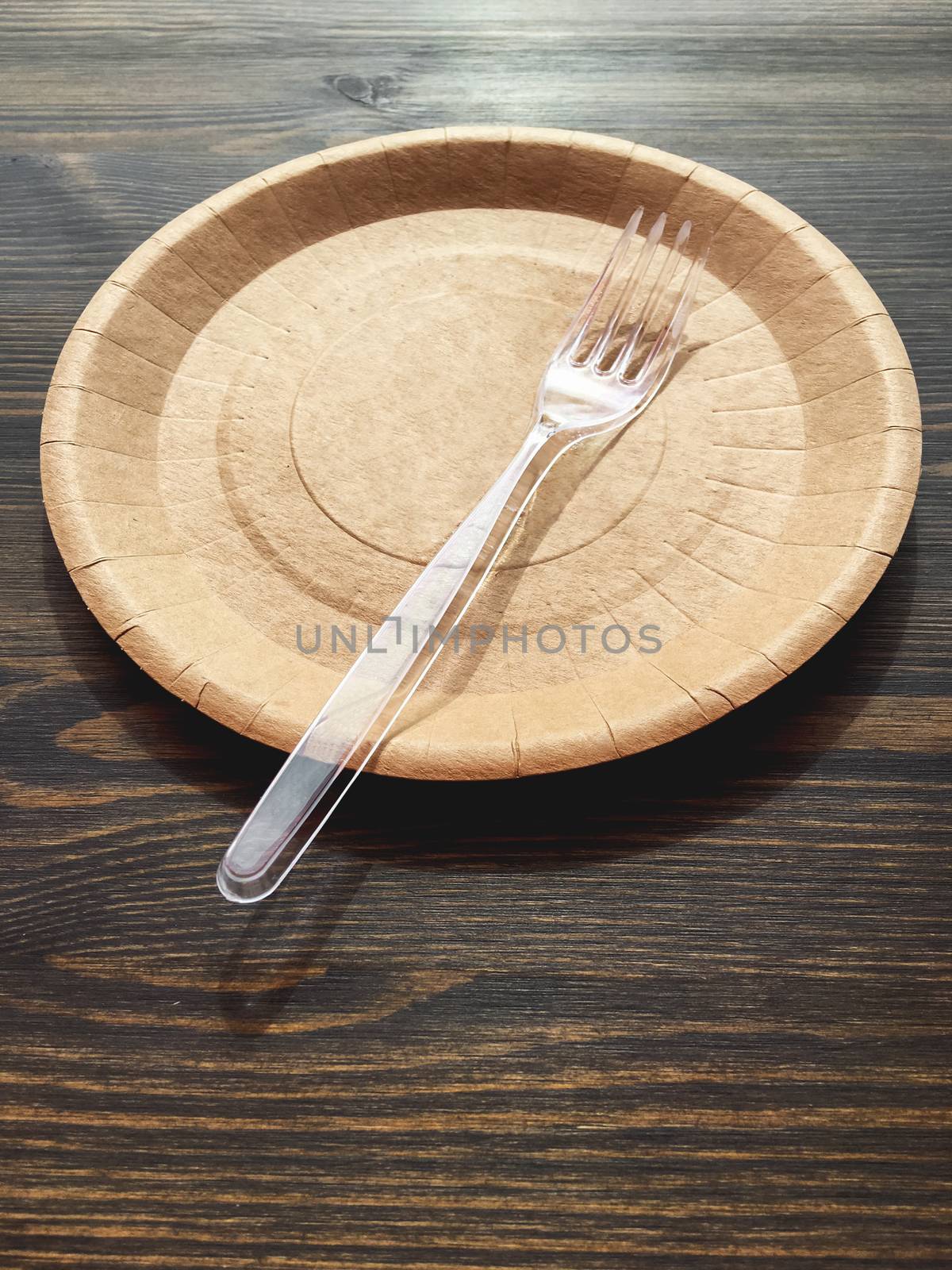Disposable tableware on wooden table in cafe. Round cardboard plate and transparent plastic fork.