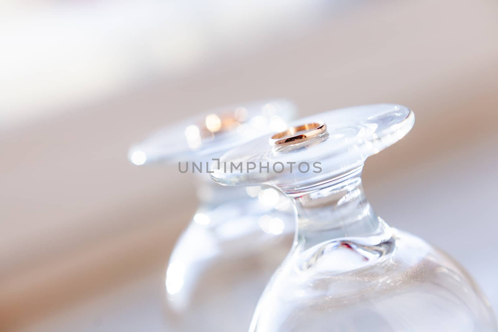 Top view on golden wedding rings lying on top of upside down staying wine glasses. Sun shines through transparent glass.