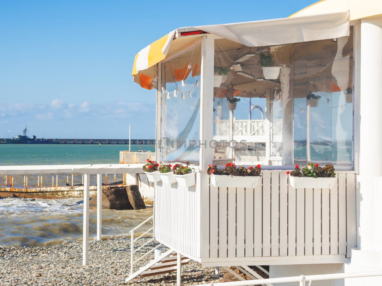 Cafe with views of Black sea and rocky beach. Balcony of white boards with flower beds at plastic windows. Sochi, Russia.