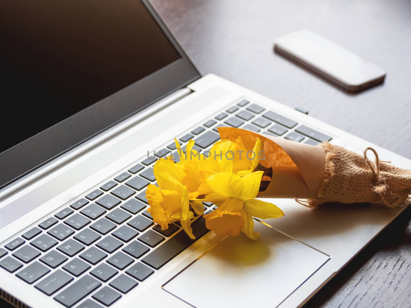 Bouquet of Narcissus or daffodils lying on silver metal laptop. Bright yellow flowers on portable device. Smartphone on wooden background.