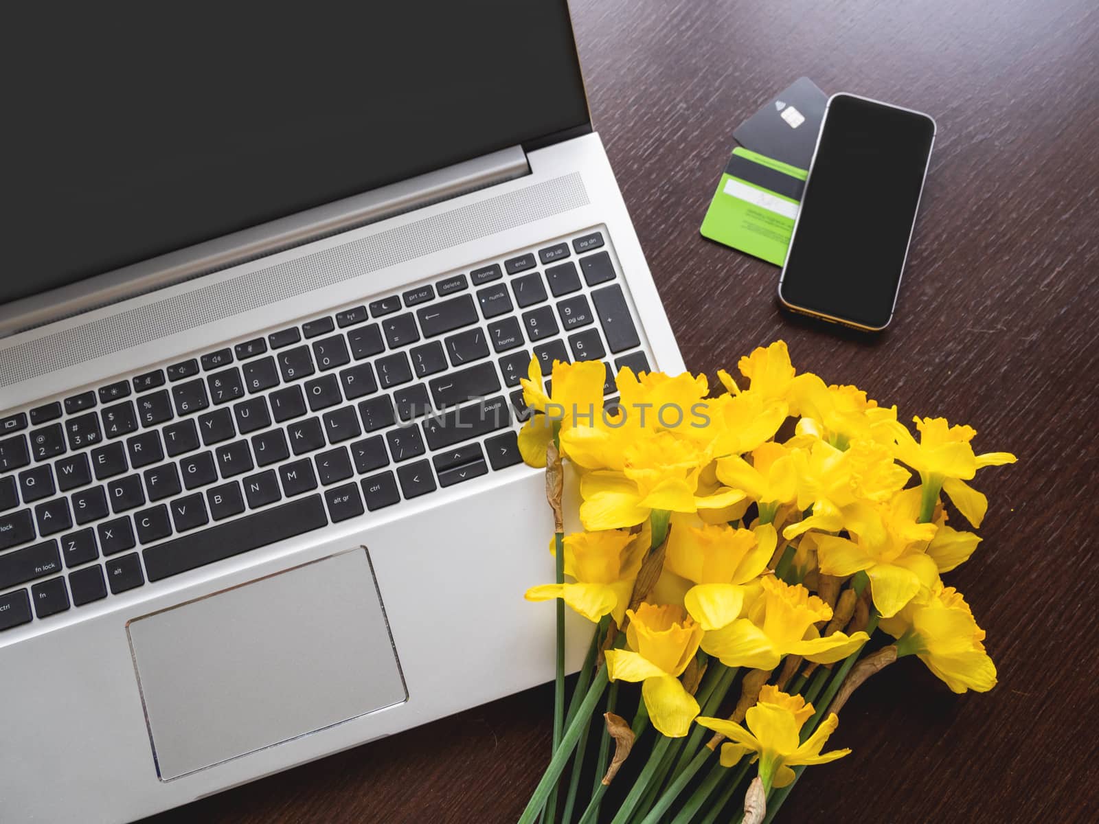 Bouquet of Narcissus or daffodils lying on silver metal laptop. Top view on bright yellow flowers on portable device and smartphone with credit cards on wooden table.