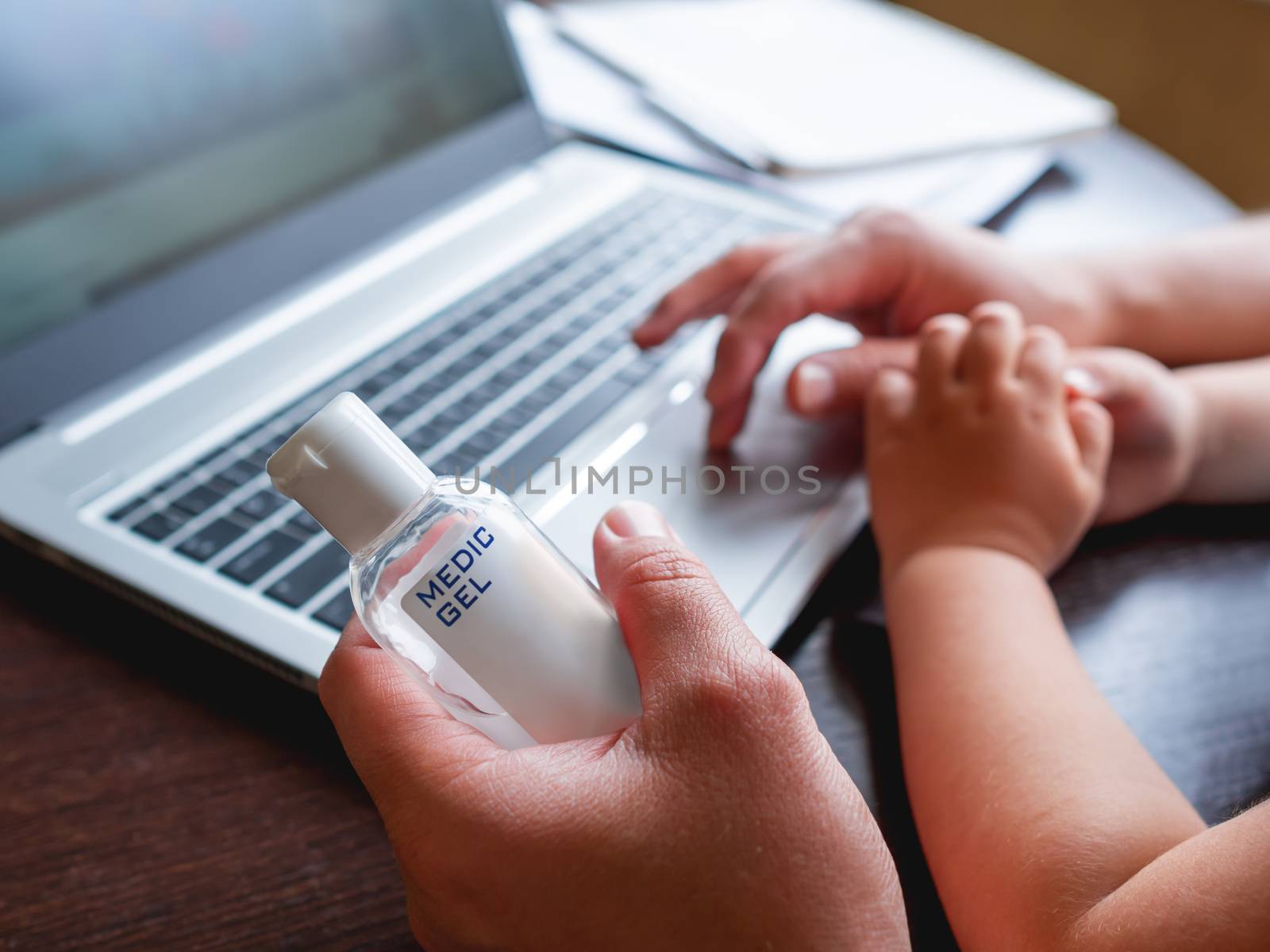 Self-isolation because of coronavirus outbreak. Hand with medical sanitizer gel for skin sterilization. Man is remote working with his little son. Laptop on table. by aksenovko