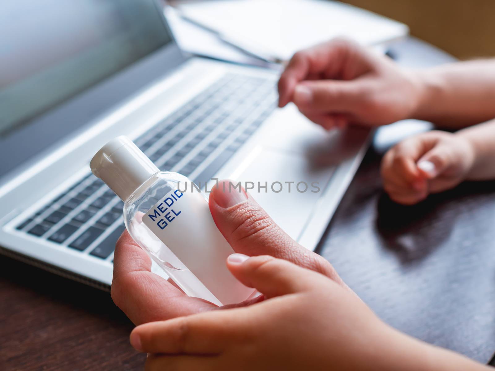 Self-isolation because of coronavirus outbreak. Hand with medical sanitizer gel for skin sterilization. Man is remote working with his little son. Laptop on table. by aksenovko