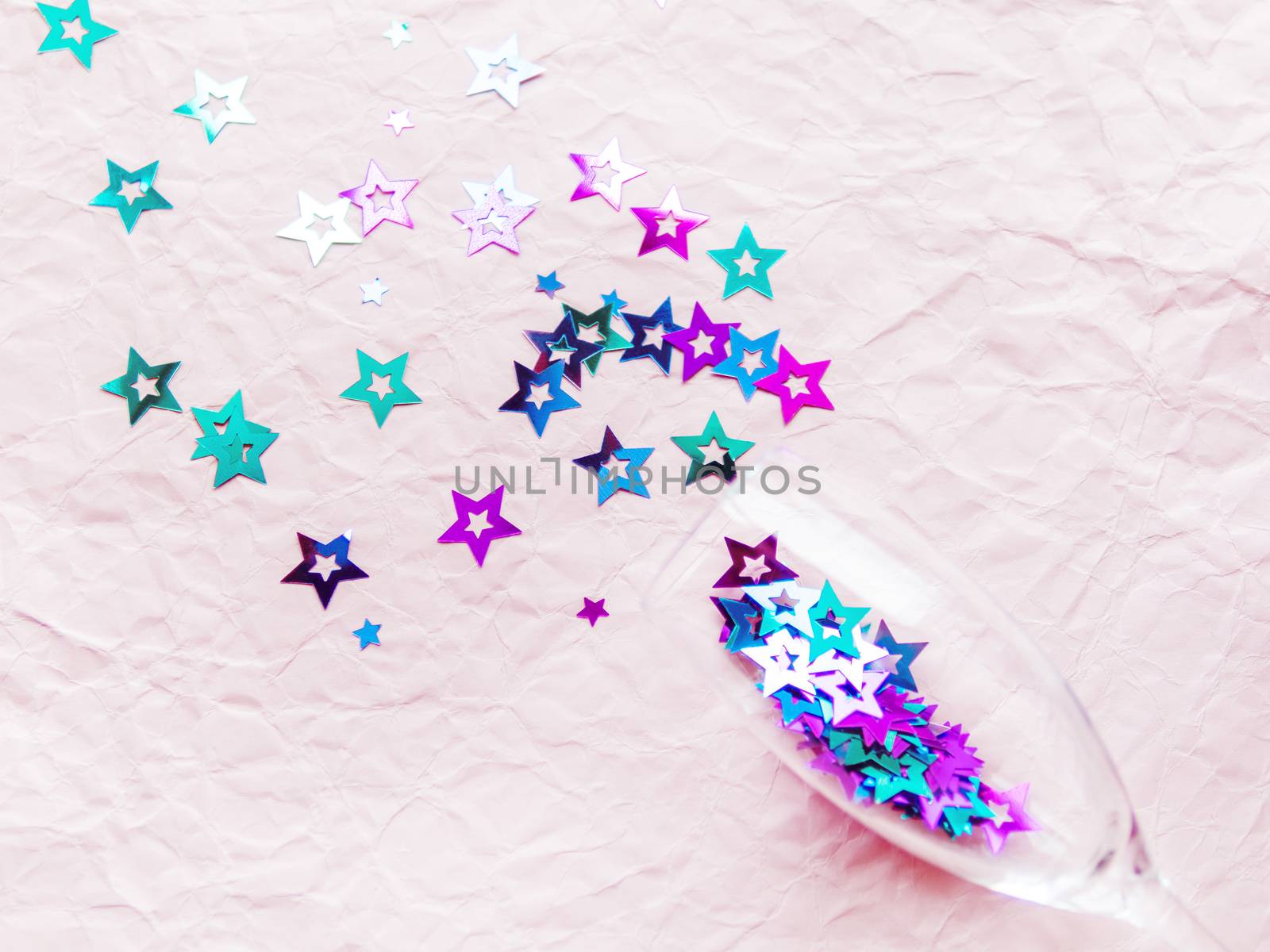 Transparent wine glass with magenta and cyan colored spangles. Festive copy space with crockery on crumpled pink paper background.