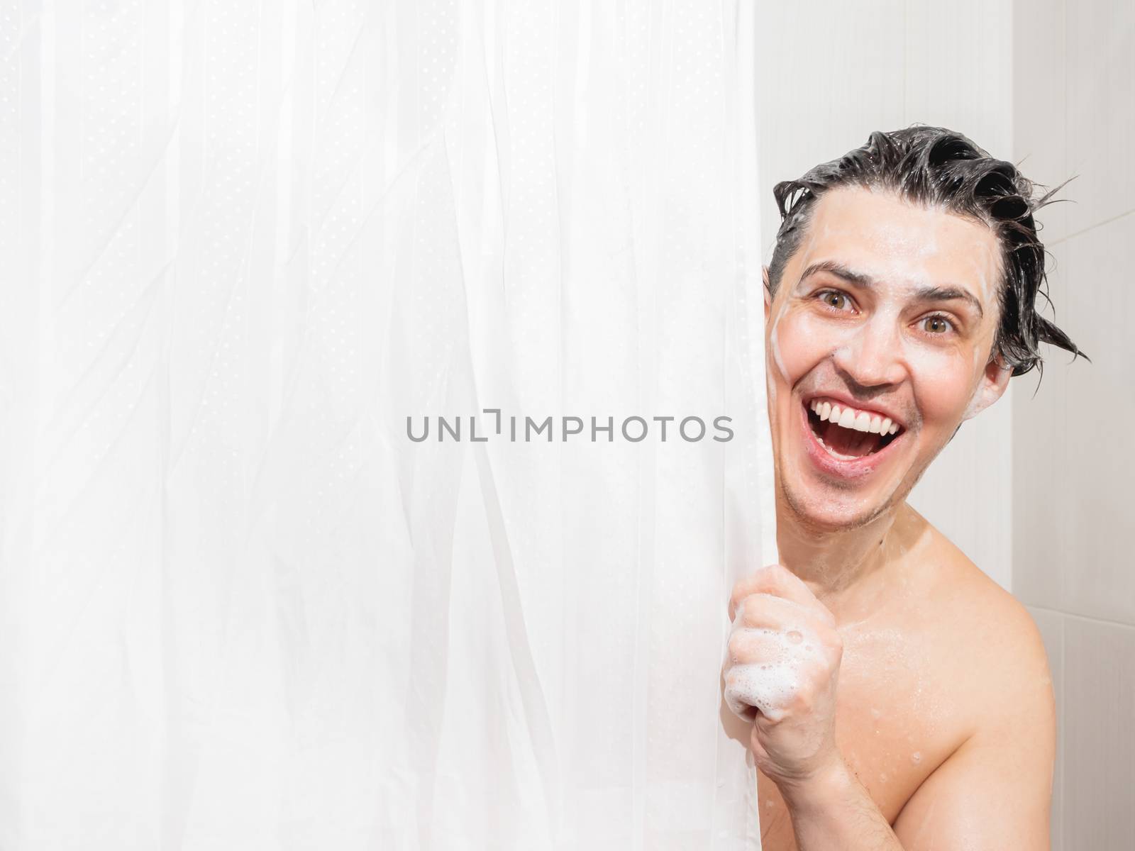 Young man in soap suds looks out from behind a curtain in the ba by aksenovko