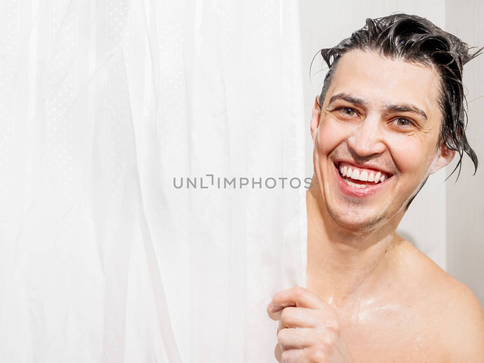 Young man in soap suds looks out from behind a curtain in the bathroom. Man was taking a shower.