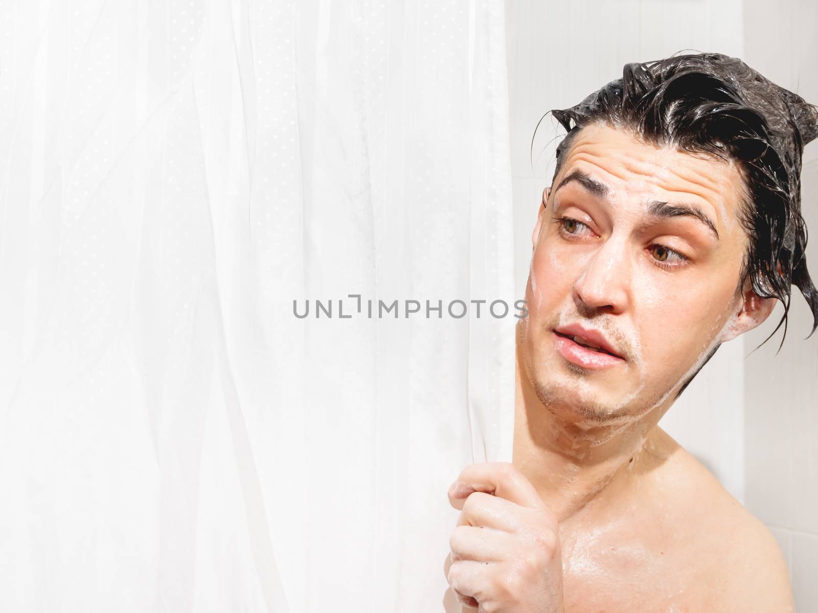 Young man in soap suds looks out from behind a curtain in the ba by aksenovko