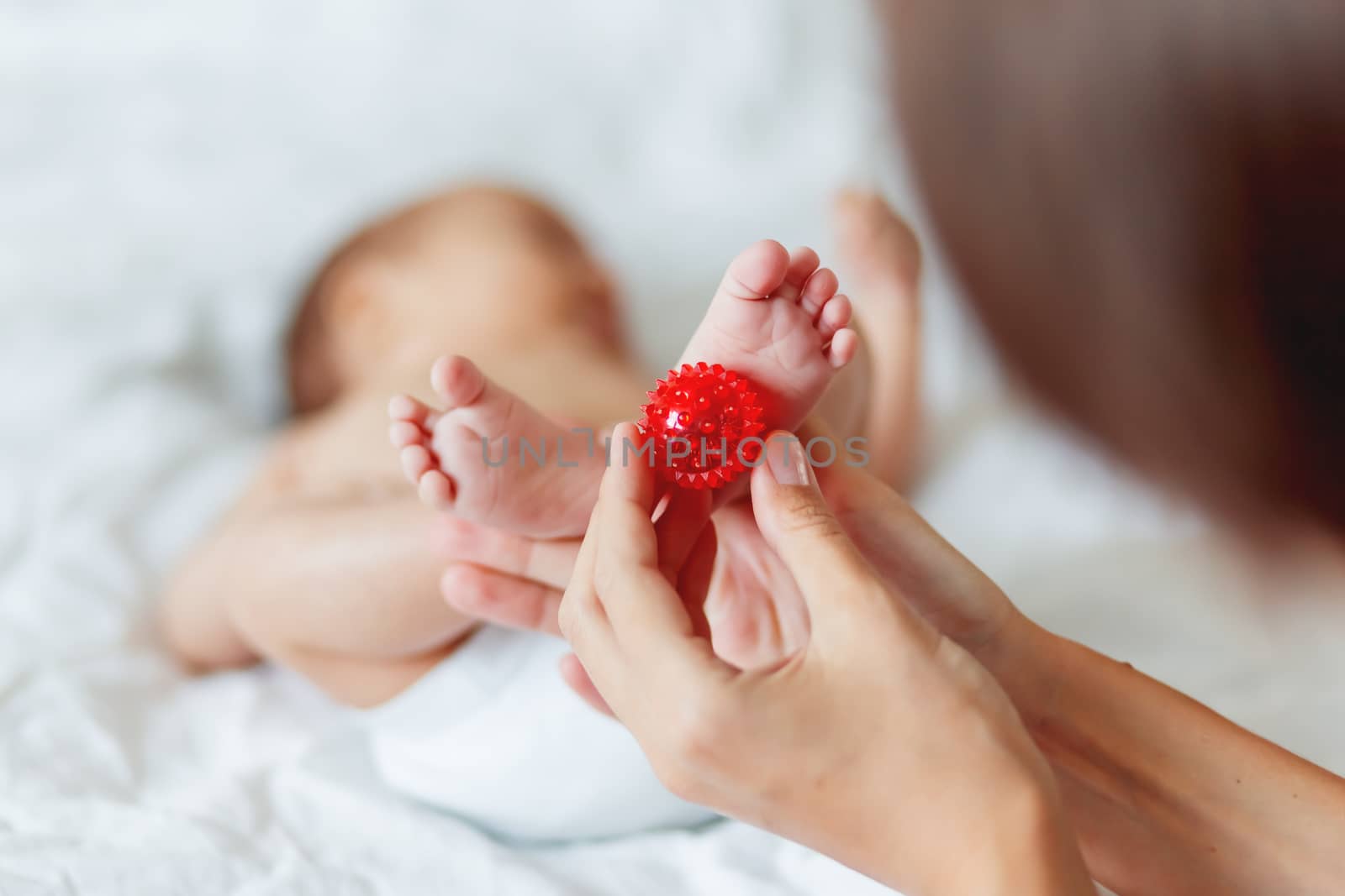 Mother holds newborn baby's feet. Tiny fingers and red massage ball in woman's hand. Cozy morning at home. by aksenovko