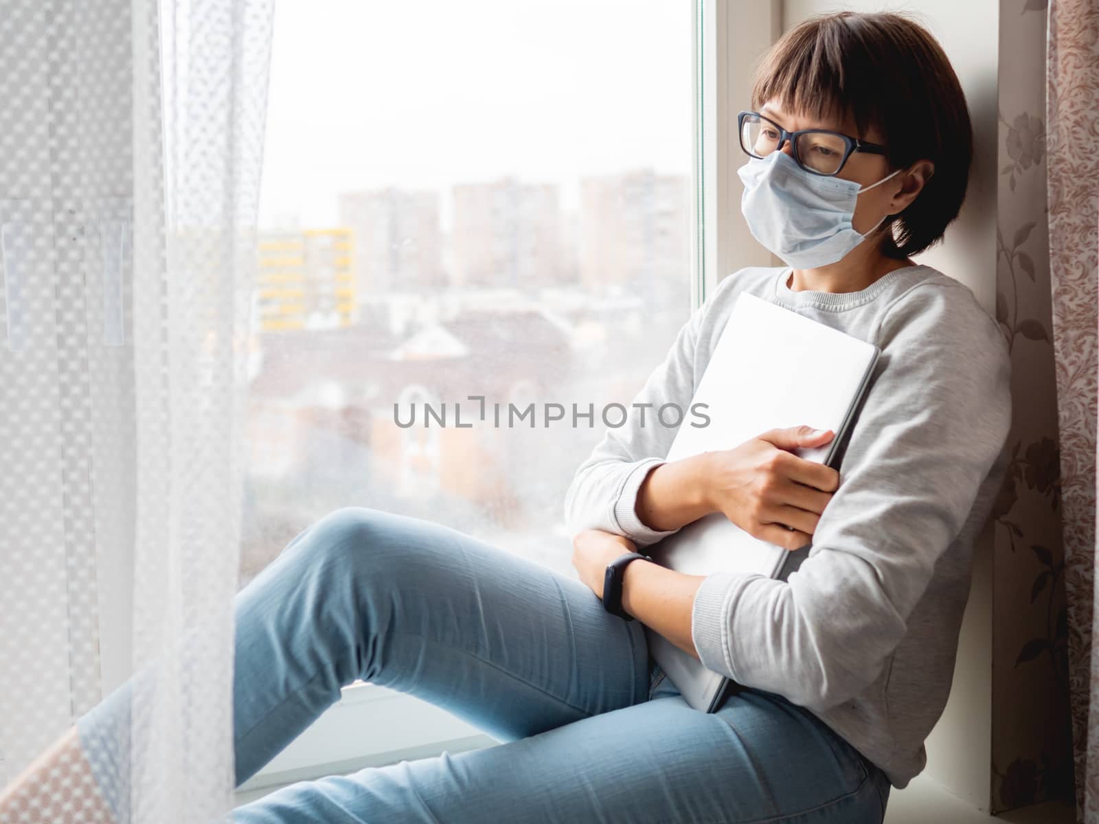 Woman in medical mask had lost her job. She sits on window sill with closed laptop. Lockdown quarantine because of coronavirus COVID19. Self isolation at home.