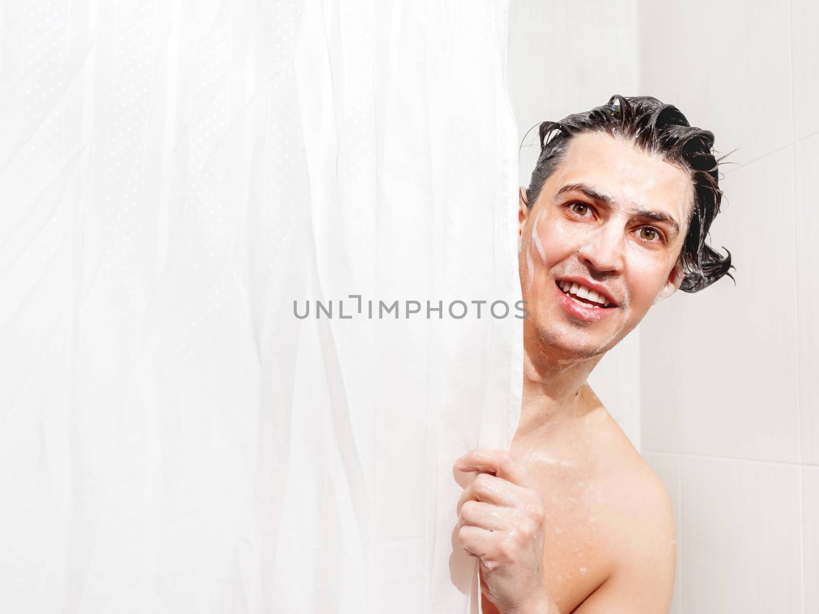 Young man in soap suds looks out from behind a curtain in the ba by aksenovko