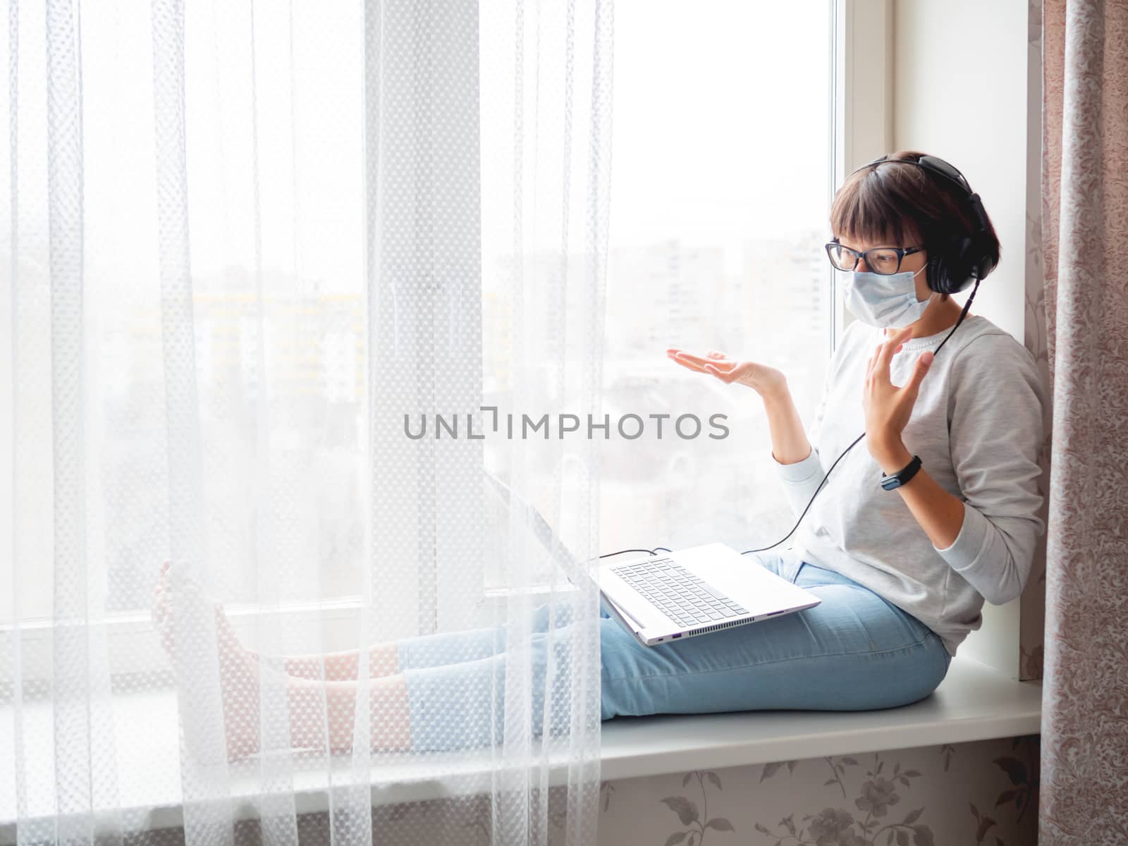 Woman in medical mask remote works from home. She speaks online with somebody on window sill with laptop on knees. Lockdown quarantine because of coronavirus COVID19. Self isolation at home.