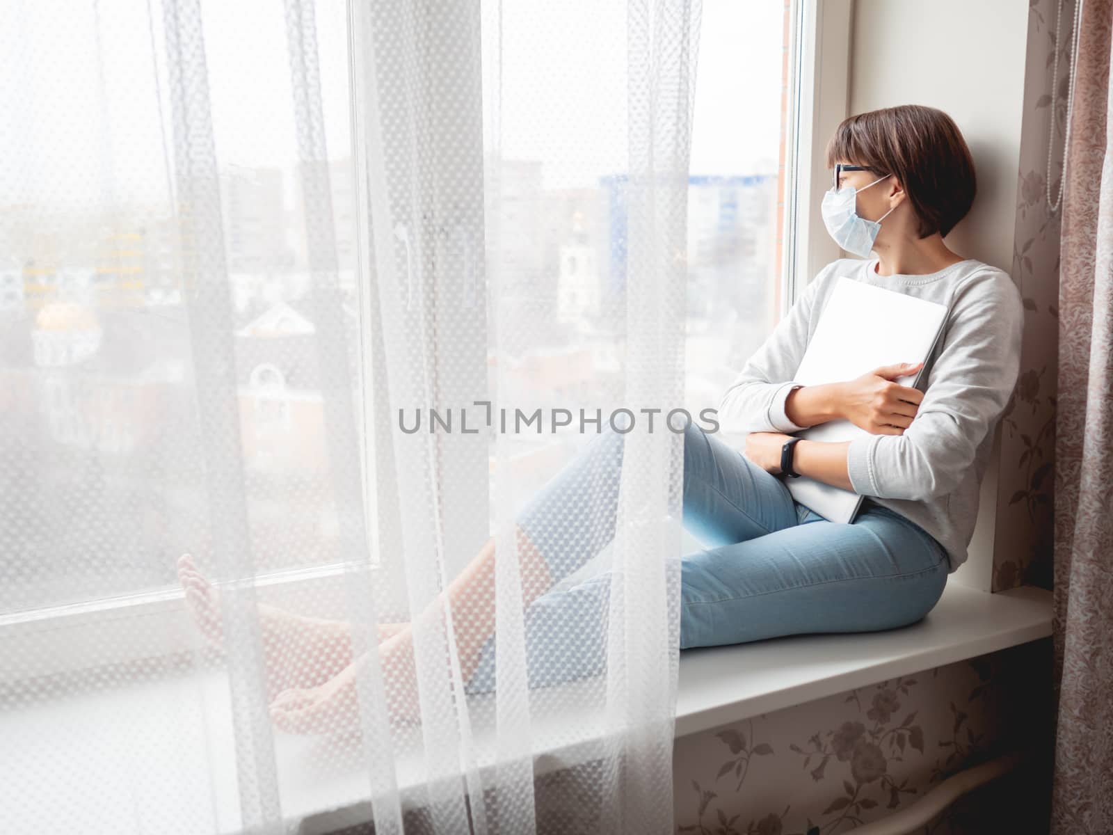 Woman in medical mask had lost her job. She sits on window sill with closed laptop. Lockdown quarantine because of coronavirus COVID19. Self isolation at home.