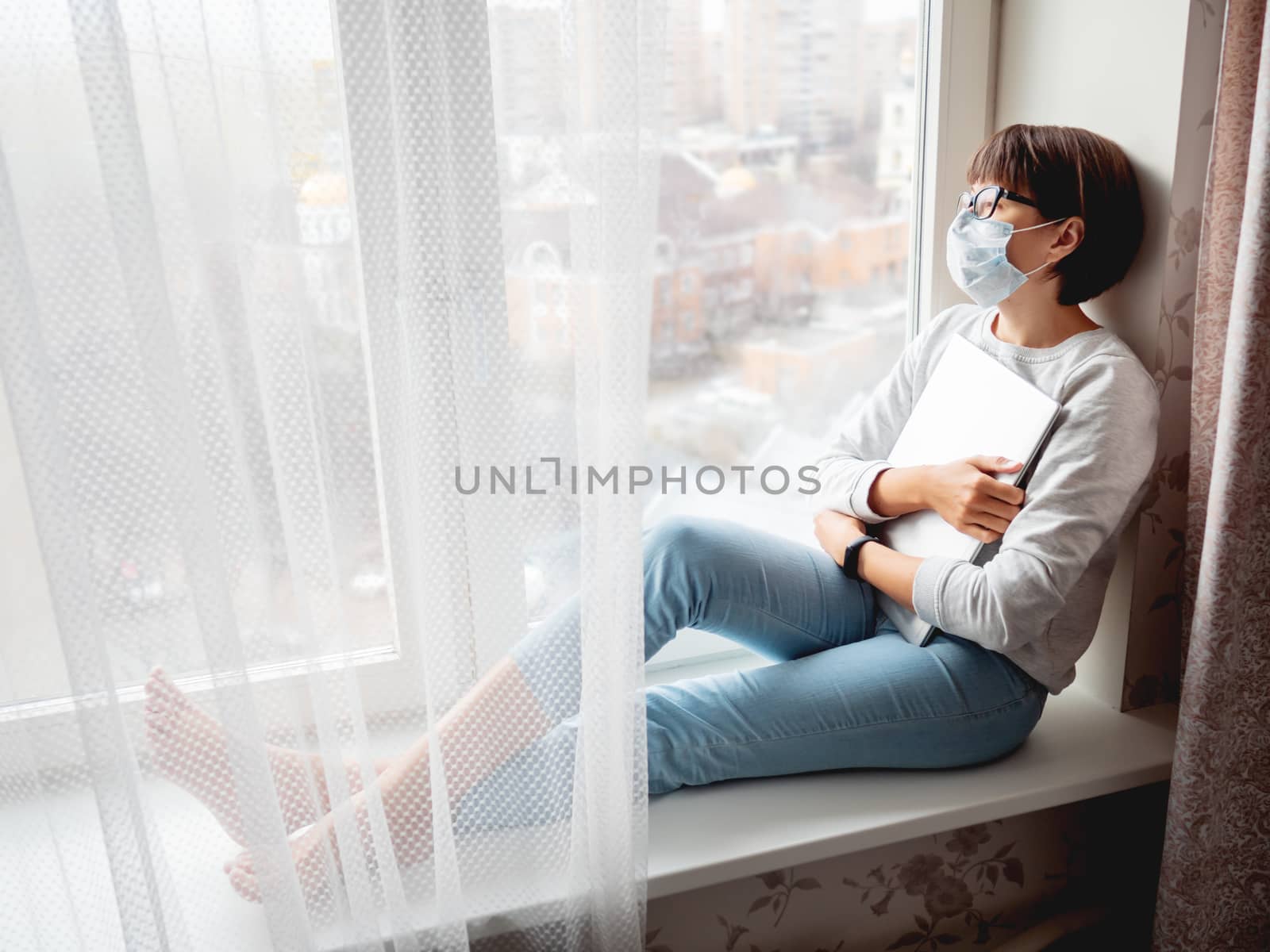 Woman in medical mask had lost her job. She sits on window sill with closed laptop. Lockdown quarantine because of coronavirus COVID19. Self isolation at home.