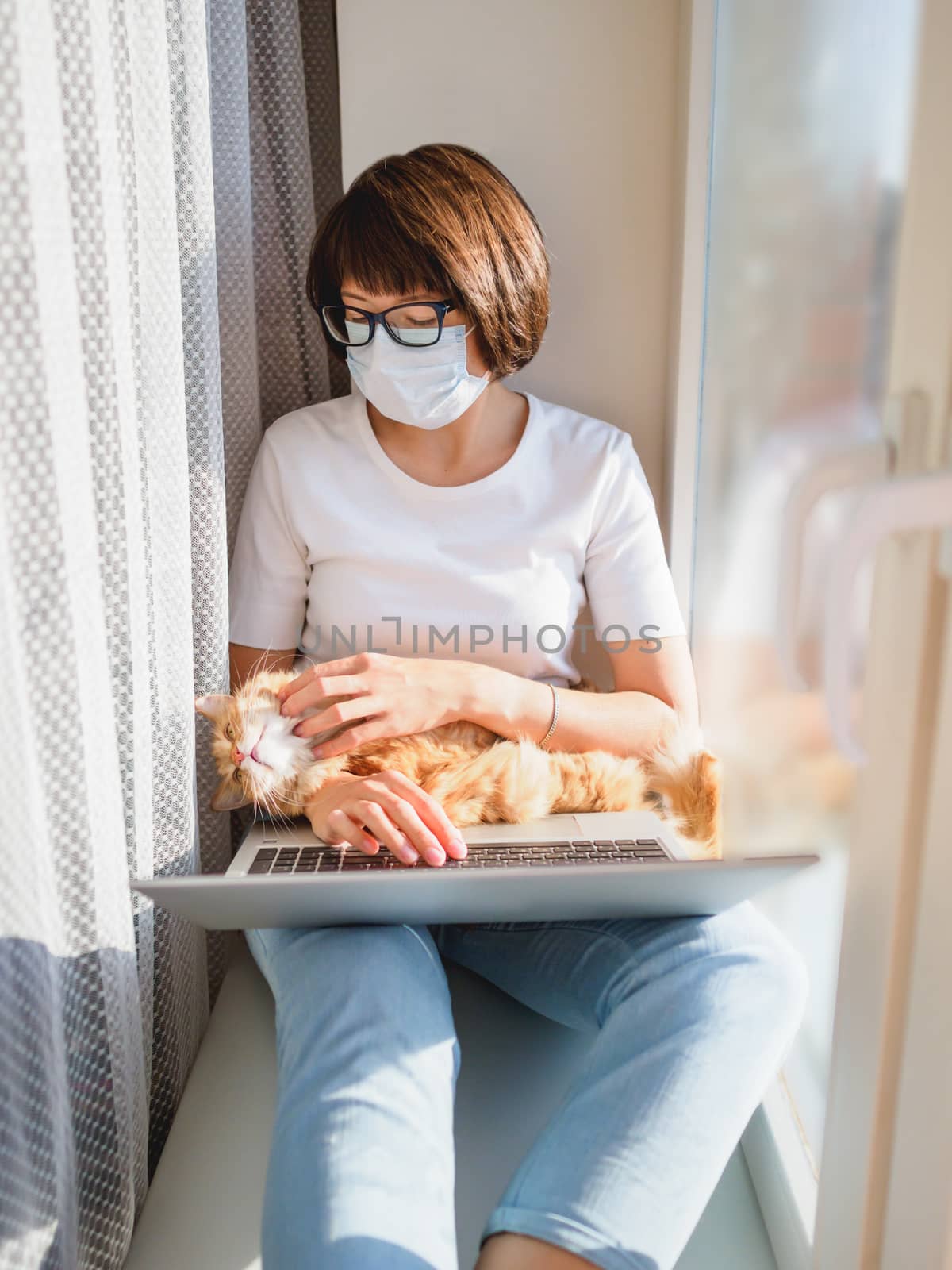 Woman in medical mask remote works from home. She sits on window sill with laptop and cute ginger cat on her knees. Lockdown quarantine because of coronavirus COVID19. Self isolation at home.