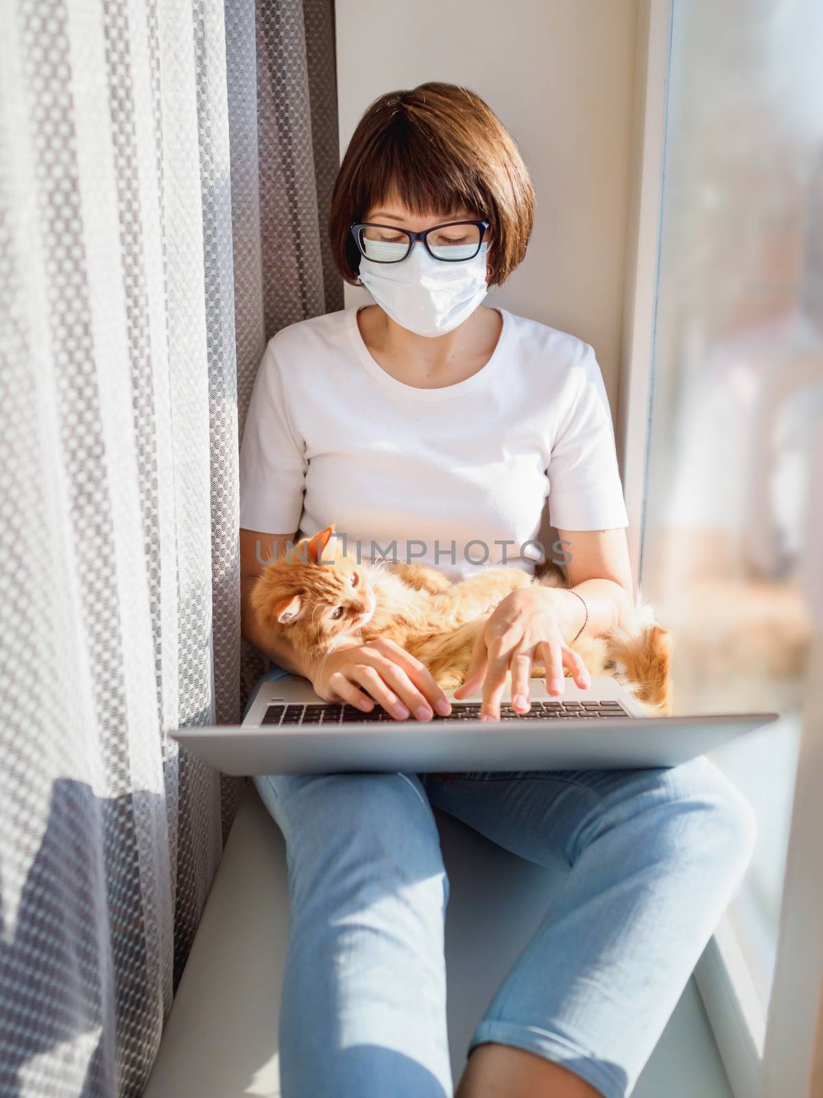 Woman in medical mask remote works from home. She sits on window sill with laptop and cute ginger cat on her knees. Lockdown quarantine because of coronavirus COVID19. Self isolation at home.