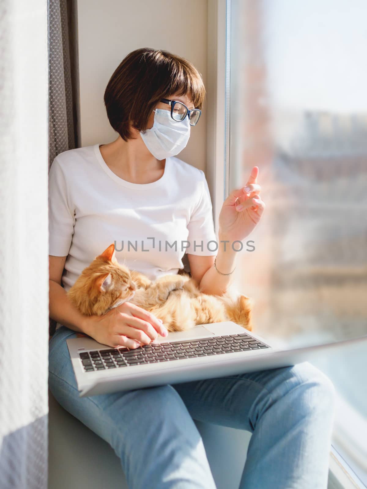 Woman in medical mask remote works from home. She sits on window sill with laptop and cute ginger cat on her knees. Lockdown quarantine because of coronavirus COVID19. Self isolation at home.