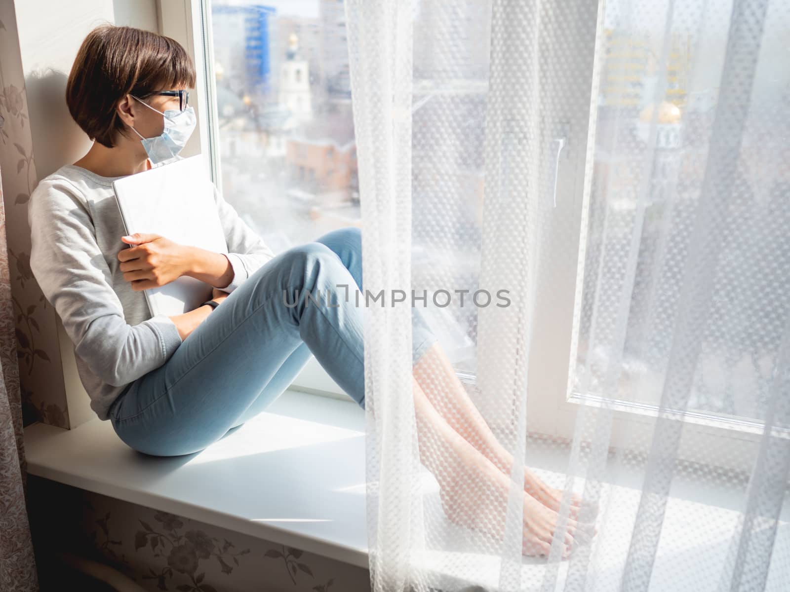 Woman in medical mask had lost her job. She sits on window sill with closed laptop. Lockdown quarantine because of coronavirus COVID19. Self isolation at home.