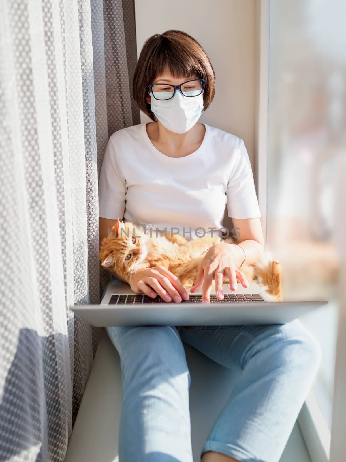 Woman in medical mask remote works from home. She sits on window by aksenovko