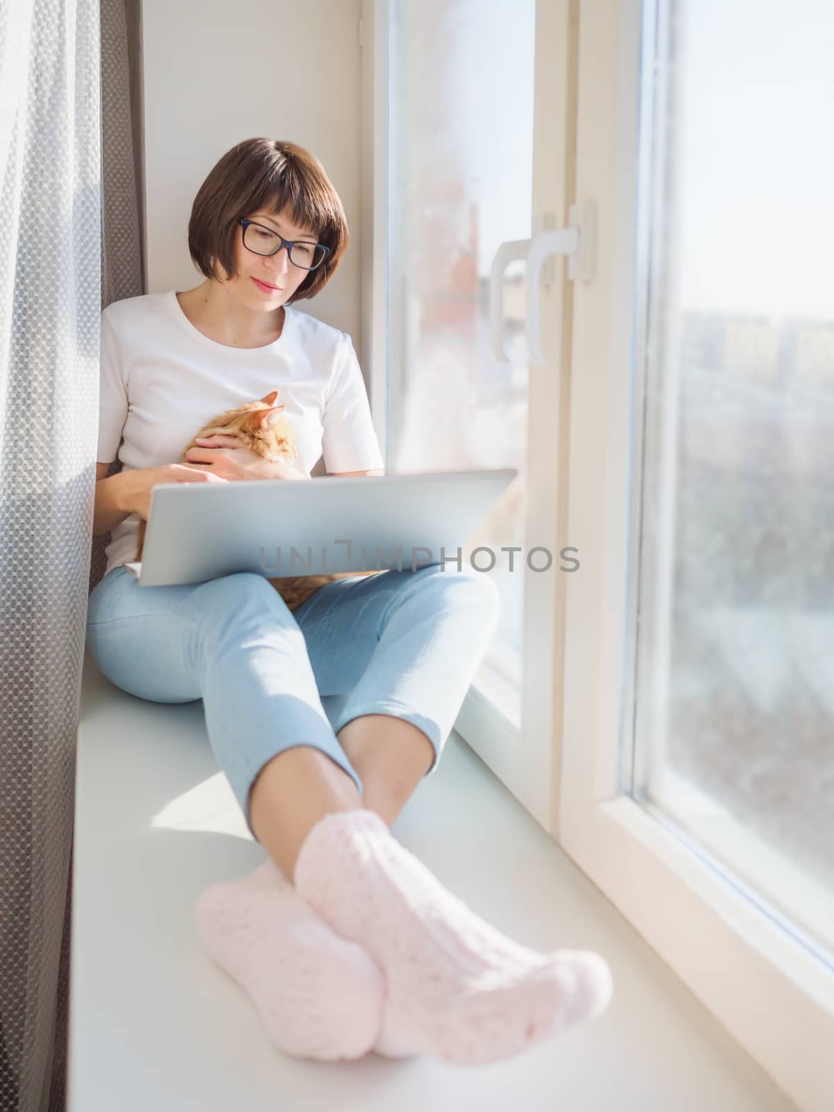 Woman remote works from home. She sits on window sill with laptop and cute ginger cat on her knees. Lockdown quarantine because of coronavirus COVID19. Self isolation at home.