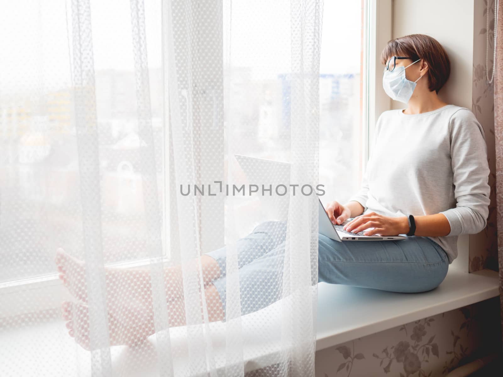 Woman in medical mask remote works from home. She sits on window by aksenovko