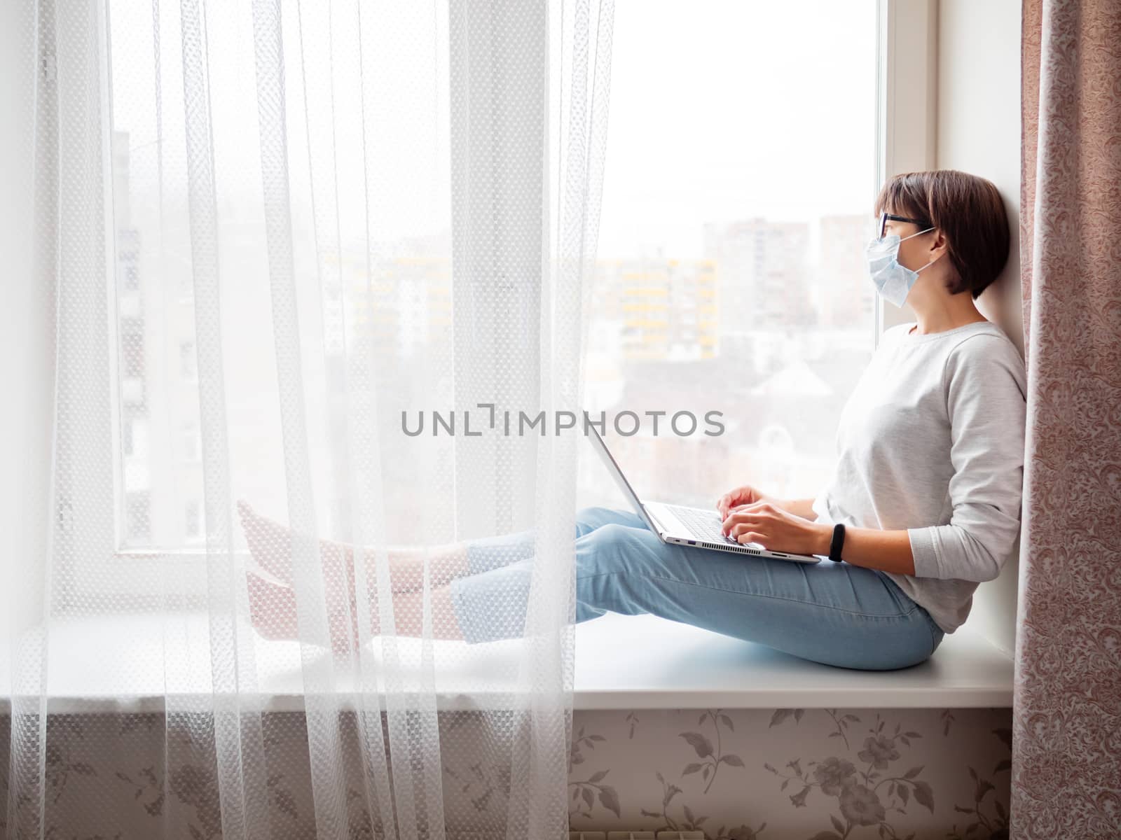 Woman in medical mask remote works from home. She sits on window sill with laptop on knees. Lockdown quarantine because of coronavirus COVID19. Self isolation at home.