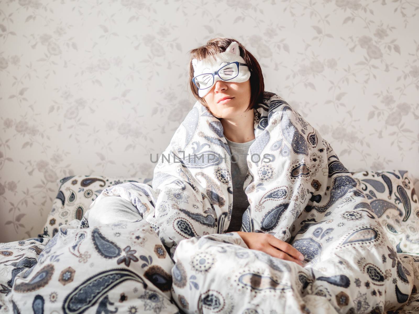 Young woman in grey pajama, glasses and sleeping mask in shape of cute sleeping cat face. She is just woke up and sit in bed. Early morning in cozy home.